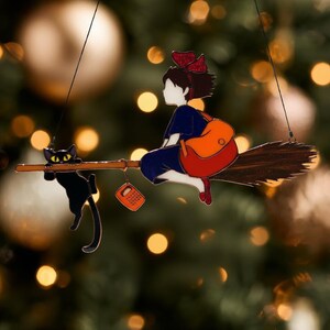 May include: A decorative ornament featuring a young witch riding a broom with a black cat. The witch wears a blue dress, red shoes, and a red bow. She carries an orange backpack. The cat clings to the broom.