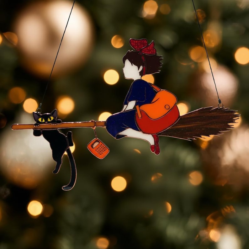 May include: A decorative ornament featuring a young witch riding a broom with a black cat. The witch wears a blue dress, red shoes, and a red bow. She carries an orange backpack. The cat clings to the broom.