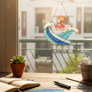 May include: A stained glass window decoration featuring a girl riding a wave. The girl wears a pink dress and has orange hair. The wave is blue and white. The decoration hangs from a suction cup on a window. A succulent in a terracotta pot sits on a wooden table.