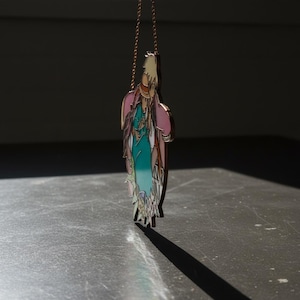 May include: A colorful pendant necklace with a unique, abstract design. The pendant features shades of pink, teal, and beige, with a gold-colored chain. The necklace is displayed on a dark gray surface, with a long shadow.