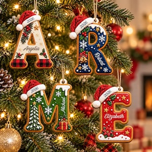 Puede incluir: Cuatro adornos navideños con forma de letra colgados en un árbol. La letra A es blanca con cuadros rojos, R es azul con copos de nieve, M es verde con un árbol y E es roja con flores de pascua. Cada uno tiene un gorro de Papá Noel, el año 2026 y un nombre.