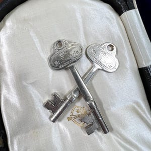 May include: Two antique silver keys with the text "St. Francis" are crossed on a white satin surface. The keys have a decorative top and a detailed key bit. The keys are inside a black case.