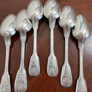 May include: Set of six silver spoons arranged on a dark wooden surface. Each spoon features a rounded bowl and a decorative handle with a shell and crest design. The spoons are made of a shiny, reflective metal.