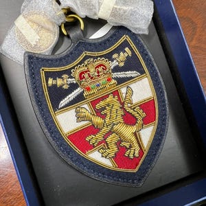 May include: A navy blue shield-shaped keychain with a gold lion and crown emblem. The emblem features red and white stripes, gold embroidery, and crossed swords. The keychain has a gold ring and a leather strap.