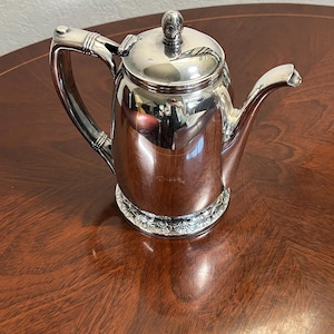 May include: A polished silver-tone metal coffee pot featuring a hinged lid with a decorative finial, a curved handle with ribbed detailing, and a flared spout. The base of the pot is adorned with a repeating embossed floral or shell pattern.