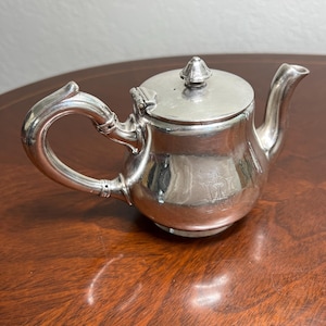 May include: A small, silver teapot with a curved handle and a spout. The lid has a decorative finial. The teapot has an engraved design on the body. The teapot is sitting on a wooden surface.