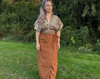 May include: A woman wearing a two-piece outfit in a natural outdoor setting. The top is a short-sleeved, wrap-style blouse with a fern pattern. The skirt is a long, wrap-style skirt in a rust-brown color.