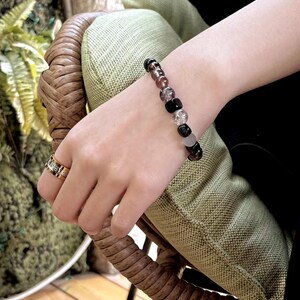 May include: A bracelet made of black, gray, and brown beads. The beads are round and have a variety of textures. The bracelet is worn on a person's wrist.