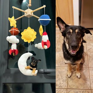 May include: A space-themed baby mobile featuring crocheted planets, stars, rockets, a sun, and a white crescent moon with a small black and tan dog figurine resting on it, suspended from a wooden frame, alongside a black and tan dog.