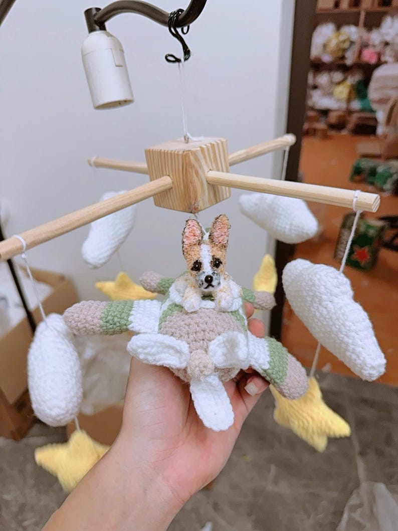 Puede incluir: Un m&oacute;vil colgante para habitaci&oacute;n infantil con un avi&oacute;n de ganchillo y una peque&ntilde;a figura de perro en la cabina. El m&oacute;vil incluye nubes blancas de ganchillo y estrellas amarillas suspendidas de un marco de madera en forma de cruz.