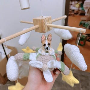 May include: A nursery mobile featuring a crocheted airplane in beige, white, and green with a small crocheted dog sitting in the cockpit. The mobile includes white crocheted clouds and yellow crocheted stars suspended from a wooden cross frame.