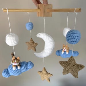 Puede incluir: Un m&oacute;vil colgante con marco de madera en cruz y elementos de ganchillo que incluyen dos gatos sobre nubes azules, una luna creciente blanca, dos estrellas beige y tres esferas blancas y azules, suspendidos por hilos blancos.