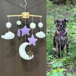 Puede incluir: Un m&oacute;vil de ganchillo para beb&eacute; con marco de madera en cruz, nubes blancas, estrellas moradas y una luna creciente blanca. Una peque&ntilde;a figura de perro de ganchillo marr&oacute;n est&aacute; unida a la luna. El m&oacute;vil cuelga de un gancho blanco.