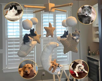 Puede incluir: Un móvil colgante para guardería con un marco de madera en forma de cruz y figuras de crochet blanco en forma de luna, estrella y esfera. Pequeñas figuras de animales de peluche están unidas a las formas. Cuatro recuadros circulares muestran un gato blanco y negro, un gato gris, un perro marrón y un bulldog blanco y negro.