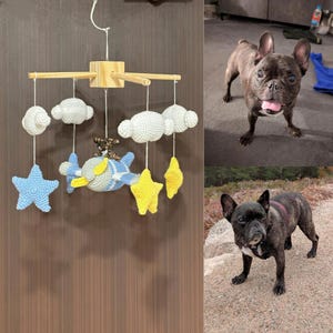Puede incluir: Un m&oacute;vil de ganchillo con marco de madera, nubes blancas, estrellas amarillas y un avi&oacute;n azul y amarillo con una peque&ntilde;a figura de perro marr&oacute;n. El m&oacute;vil est&aacute; suspendido por un hilo blanco sobre una superficie vertical marr&oacute;n oscuro.