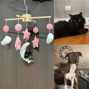 Puede incluir: Un m&oacute;vil de madera con elementos de ganchillo que incluyen esferas rosas, estrellas rosas, nubes blancas y una luna creciente blanca. Un peque&ntilde;o gato negro y un perro peque&ntilde;o marr&oacute;n y blanco est&aacute;n unidos a la luna. El m&oacute;vil cuelga de un bloque central.