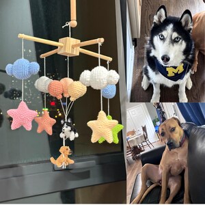 May include: A wooden mobile featuring crocheted clouds, stars, and two small dog figures hanging from white strings. The mobile includes light blue, white, peach, yellow, pink, and green yarn elements suspended from a central wooden cross frame.