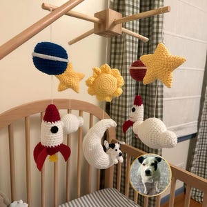 Puede incluir: Un m&oacute;vil de cuna con tem&aacute;tica espacial que incluye elementos de ganchillo como un planeta azul, estrellas amarillas, un sol amarillo, un cohete rojo y blanco, una luna creciente blanca y un perro peque&ntilde;o, suspendidos de un marco de madera.