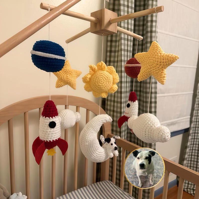 Puede incluir: Un m&oacute;vil de cuna con tem&aacute;tica espacial que incluye elementos de ganchillo como un planeta azul, estrellas amarillas, un sol amarillo, un cohete rojo y blanco, una luna creciente blanca y un perro peque&ntilde;o, suspendidos de un marco de madera.