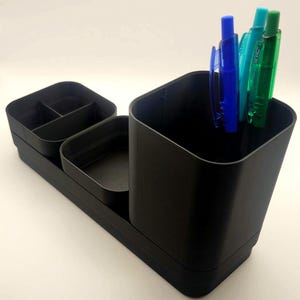 May include: A black desk organizer with multiple compartments. It includes a divided tray, a small square container, and a larger square cup. Several pens in blue and green are placed in the cup.