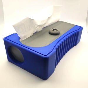 Könnte beinhalten: Eine blaue und graue Taschentuchbox in Form eines Bleistiftanspitzers. Die Box hat eine graue Oberseite mit einem grauen Schraubendetail und eine blaue Basis. Weiße Taschentücher ragen aus der oberen Öffnung heraus.