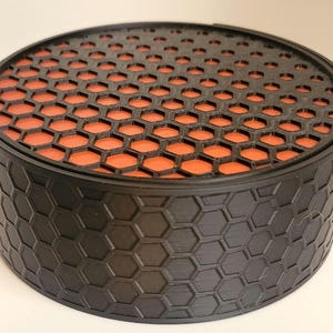 May include: A black round container with a honeycomb pattern on the outside and a red honeycomb pattern on the lid.