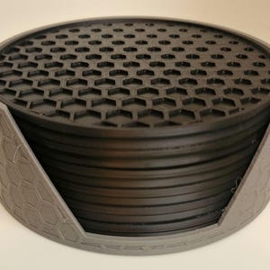 May include: A set of black hexagonal coasters with a grey holder. The coasters are stacked on top of each other and the holder has a hexagonal pattern.