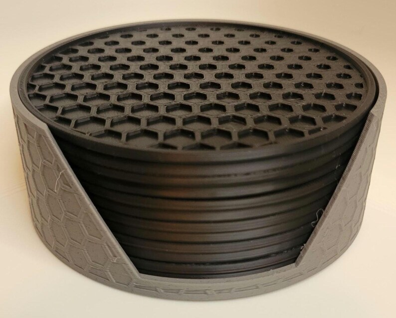 May include: A set of black hexagonal coasters with a grey holder. The coasters are stacked on top of each other and the holder has a hexagonal pattern.