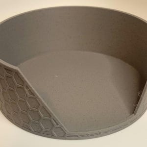 May include: A grey ceramic pet food bowl with a honeycomb pattern on the outside. The bowl has a raised lip around the edge and a slightly angled opening.