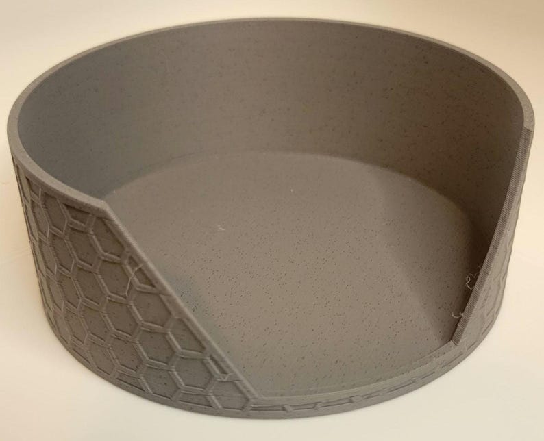 May include: A grey ceramic pet food bowl with a honeycomb pattern on the outside. The bowl has a raised lip around the edge and a slightly angled opening.