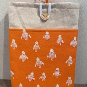 May include: A rectangular tote bag featuring a bright orange front panel decorated with a repeating pattern of white and pink sea turtles. The top section is a natural beige colour. A wooden button and a metal ring are attached.
