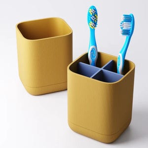 May include: Mustard yellow toothbrush holder with a ribbed texture, divided into four sections with a blue interior. Two blue toothbrushes with white and blue bristles are in the holder. A second, matching holder is next to it.