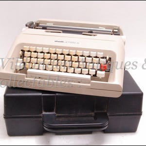 May include: A vintage Olivetti Lettera 35 typewriter in beige, with a full keyboard of off-white keys and a red key. It sits on a black carrying case with a handle. The typewriter is a classic office supply.