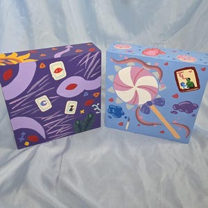 May include: Two painted boxes with colorful designs. One box is purple with white and yellow abstract shapes and symbols. The other is light blue with a large pink and white lollipop design and other small illustrations.
