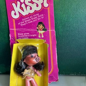 May include: A vintage Ideal Kissy doll in its original packaging. The doll has dark hair, prominent red lips, and wears a gold-toned dress. The pink cardboard backing features the text Kissy, Collect all three of the Sweet Kissy Dolls, and Kissy gives hundreds of bright red kisses.