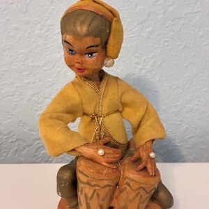 May include: A vintage figurine of a person playing the bongo drums. The figure is dressed in a yellow outfit with a matching head covering. The bongo drums are brown with a light brown base. The figure has a pearl earring and a pearl on each hand.
