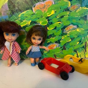 May include: Two small dolls with brown hair and blue eyes, one wearing a red and white striped coat and the other in a blue and white checkered dress, accompanied by a small red plastic toy car and a yellow plastic toy car body.