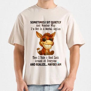 Puede incluir: Camiseta beige con un burro de dibujos animados con gafas de sol y una gran sonrisa. La camiseta tiene el texto: "SOMETIMES I SIT QUIETLY And Wonder Why I'm Not In A Mental Asylum Then I Take A Good Look Around At Everyone AND REALIZE... MAYBE I AM."