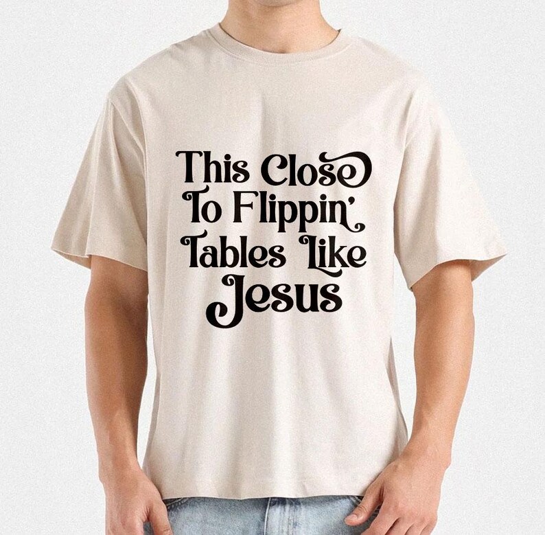 May include: A beige t-shirt with the text "This Close To Flippin' Tables Like Jesus" in a black, decorative font. The shirt has short sleeves and a relaxed fit.