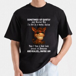Puede incluir: Camiseta negra con un burro de dibujos animados con gafas de sol y una gran sonrisa. La camiseta tiene el texto: "SOMETIMES I SIT QUIETLY And Wonder Why I'm Not In A Mental Asylum Then I Take A Good Look Around At Everyone AND REALIZE... MAYBE I AM."