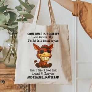 Puede incluir: Bolso tote de lona beige con un dibujo humor&iacute;stico de un burro sonriente con gafas de sol. El texto dice: "A veces me siento en silencio y me pregunto por qu&eacute; no estoy en un manicomio. Luego miro a mi alrededor... tal vez lo est&eacute;."