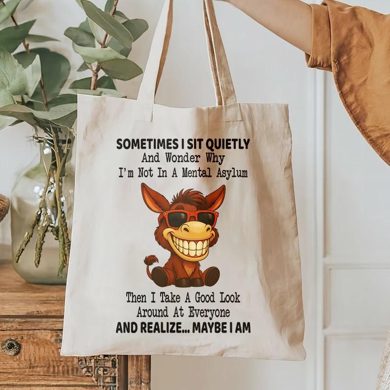 Puede incluir: Bolso tote de lona beige con un dibujo humor&iacute;stico de un burro sonriente con gafas de sol. El texto dice: "A veces me siento en silencio y me pregunto por qu&eacute; no estoy en un manicomio. Luego miro a mi alrededor... tal vez lo est&eacute;."