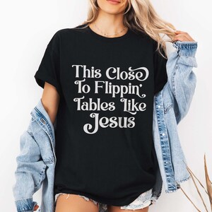 May include: Black t-shirt with the white text "This Close To Flippin' Tables Like Jesus". The shirt has short sleeves and a relaxed fit. The person is wearing a denim jacket over the shirt.