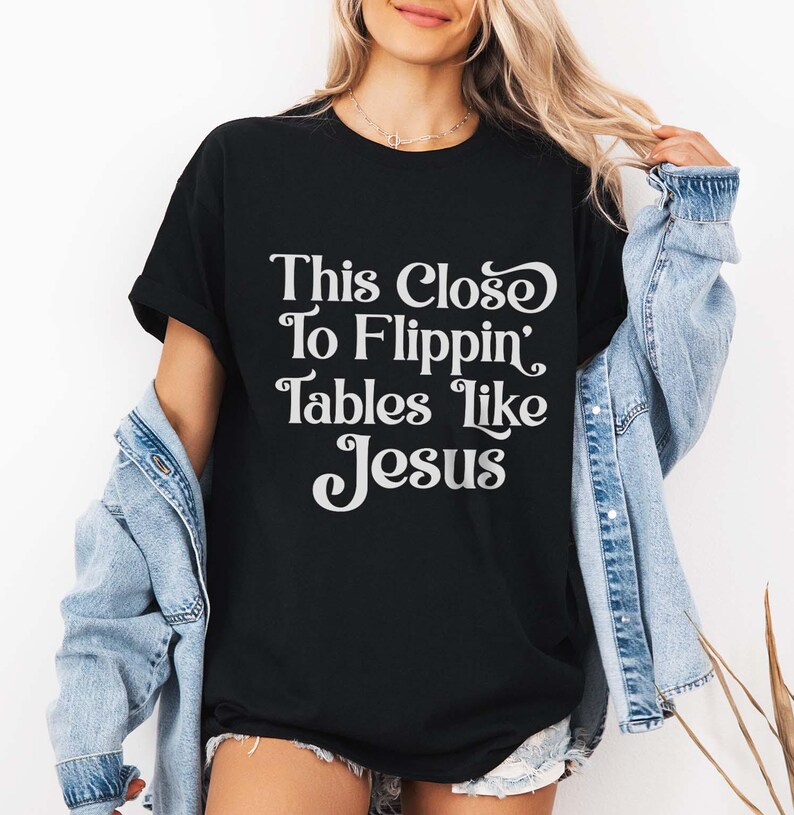 May include: Black t-shirt with the white text "This Close To Flippin' Tables Like Jesus". The shirt has short sleeves and a relaxed fit. The person is wearing a denim jacket over the shirt.
