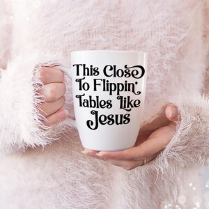 May include: White ceramic mug with the text "This Close To Flippin' Tables Like Jesus" in black script. The mug is held by a person wearing a fluffy, light pink sweater.