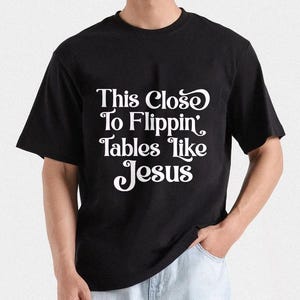 May include: Black t-shirt with the white text "This Close To Flippin' Tables like Jesus". The shirt has short sleeves and a crew neck. The person is wearing light blue jeans.
