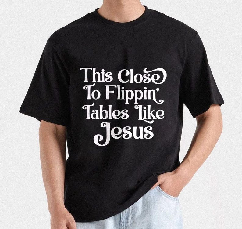 May include: Black t-shirt with the white text "This Close To Flippin' Tables like Jesus". The shirt has short sleeves and a crew neck. The person is wearing light blue jeans.