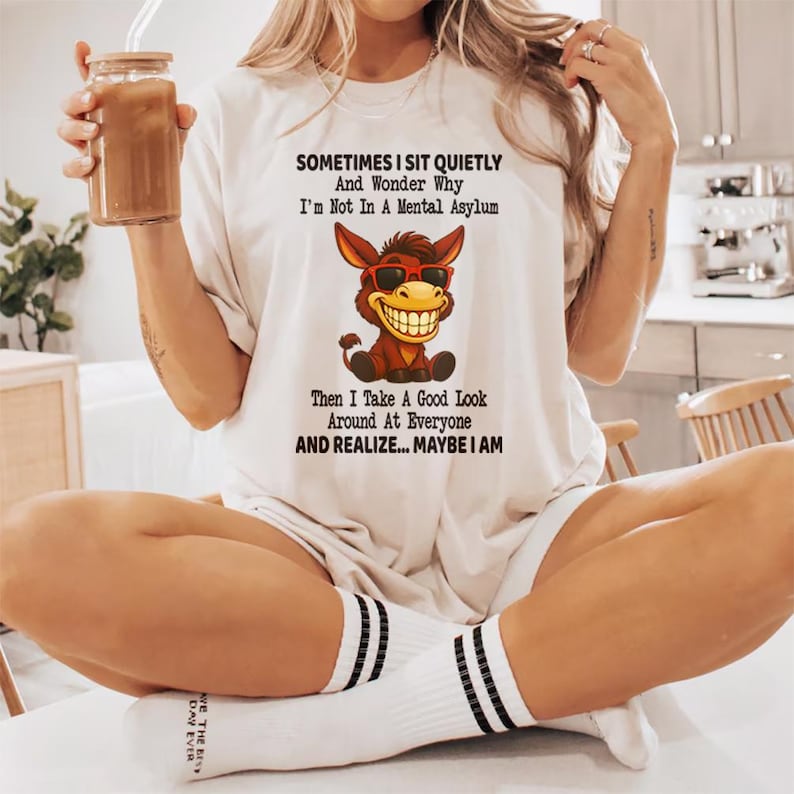 Puede incluir: Camiseta color crema con el texto "SOMETIMES I SIT QUIETLY And Wonder Why I'm Not In A Mental Asylum Then I Take A Good Look Around At Everyone AND REALIZE... MAYBE I AM." La camiseta presenta un burro de dibujos animados con gafas de sol.
