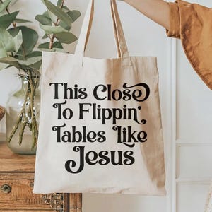 May include: A beige canvas tote bag with the phrase "This Close To Flippin' Tables Like Jesus" printed in black. The bag has two handles and is being held up by a person.