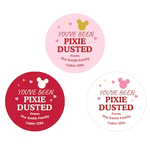 May include: Three circular stickers in light pink, dark red, and white. Each features the text "YOU'VE BEEN PIXIE DUSTED" with a mouse head silhouette, small hearts, and stars. Below, the text reads "From: The Smith Family" and "Cabin 1234".